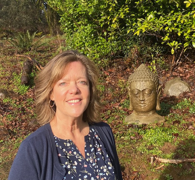 A woman with shoulder-length hair stands smiling in a garden. Shes wearing a navy blue cardigan over a floral blouse. Beside her is a stone Buddha head statue. The background is lush with green foliage and sunlight filtering through.