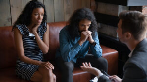 A couple sits on a brown couch in a serious conversation with a man in a suit. The woman wears a striped dress, and the man has long curly hair and a checkered shirt. The setting is a cozy room with wood and brick elements.