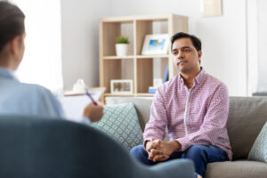 A man in a plaid shirt sits on a couch, attentively listening to someone off-camera holding a clipboard. The setting appears to be a cozy living room or office, with shelves and decor in the background.