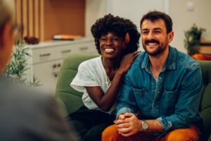 A smiling couple, one in a white shirt and the other in a denim shirt, sit on a green couch during a conversation. They are facing a blurred person in the foreground. The setting appears to be a cozy living room.