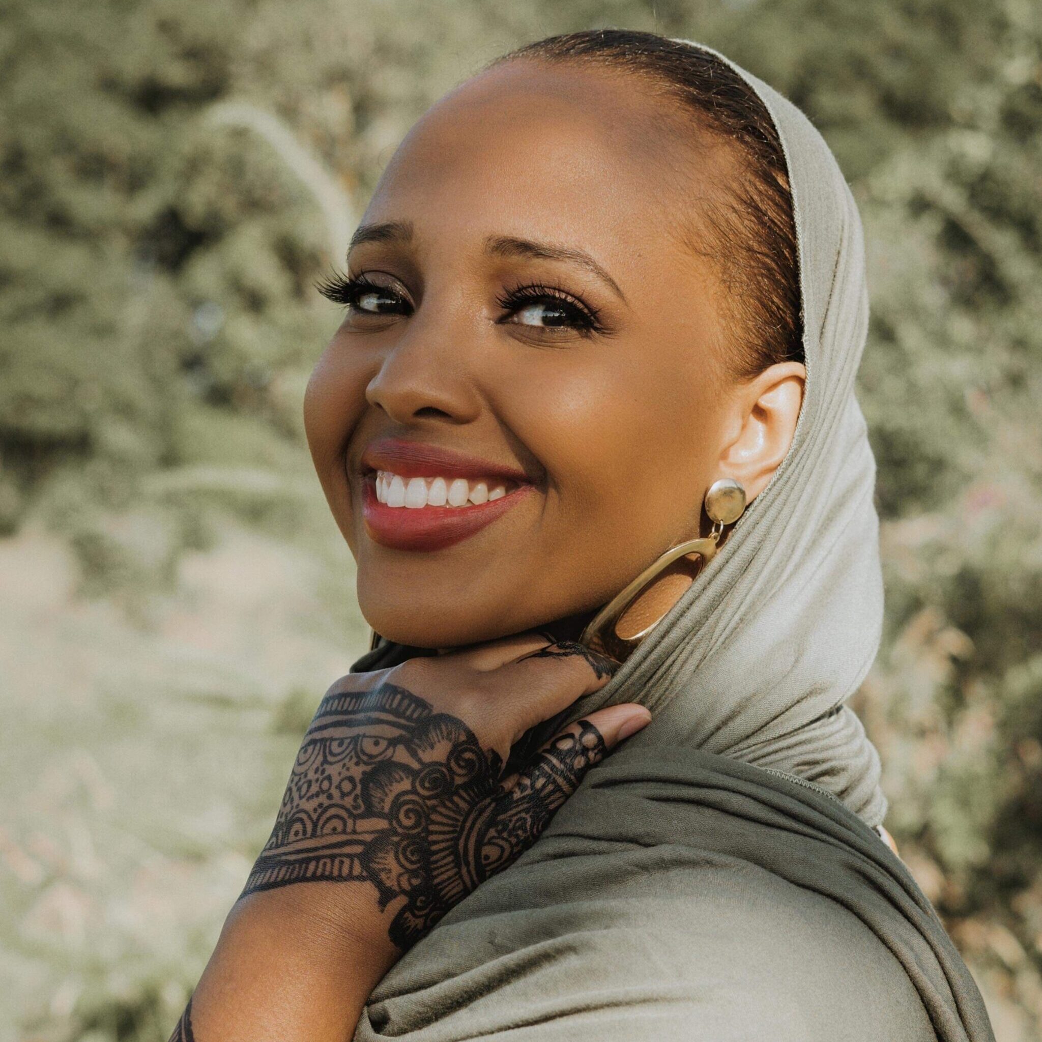 A woman wearing a light gray headscarf smiles brightly outdoors. She has intricate black henna designs on her hand and wrist, and is dressed modestly. Trees and greenery are softly blurred in the background.