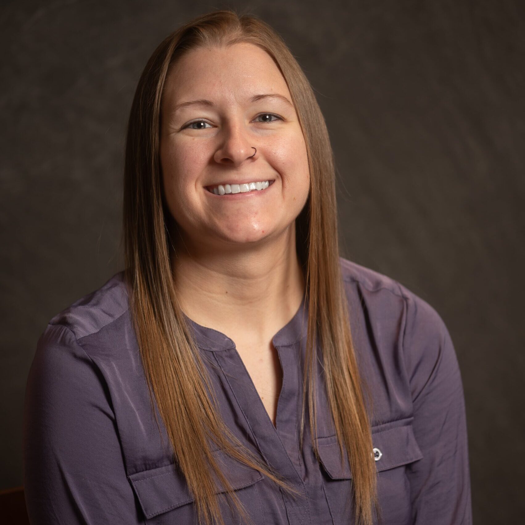 A woman with straight, light brown hair and a nose ring smiles at the camera. She is wearing a long-sleeved, button-up, purple blouse and is posed in front of a dark, neutral background.