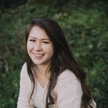 A young woman with long brown hair, wearing a cream-colored sweater and blue jeans, is kneeling outdoors on green grass and smiling at the camera. The background is blurred with greenery.