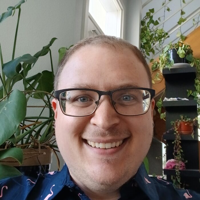 A smiling person with glasses and a flamingo-patterned shirt takes a selfie indoors, surrounded by various green potted plants near a window and staircase.