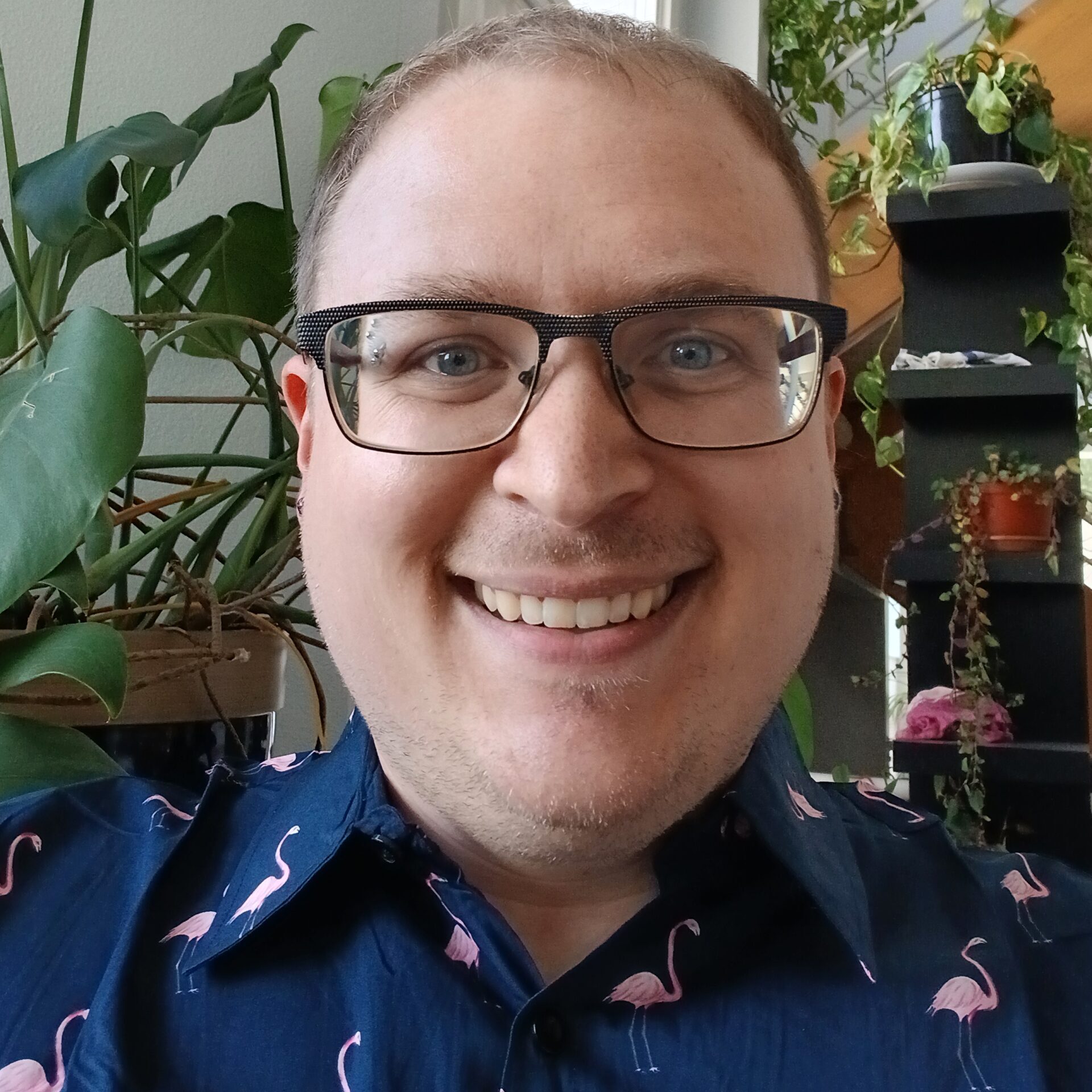 A smiling person with glasses and a flamingo-patterned shirt takes a selfie indoors, surrounded by various green potted plants near a window and staircase.