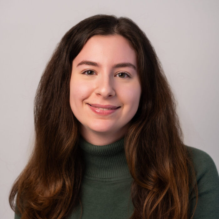 A young woman with long, wavy brown hair and a green turtleneck sweater smiles softly at the camera against a plain light gray background.