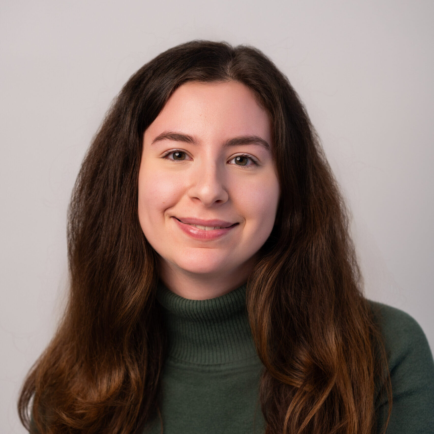 A young woman with long, wavy brown hair and a green turtleneck sweater smiles softly at the camera against a plain light gray background.