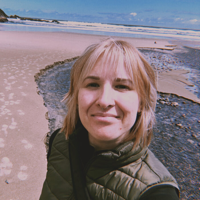 A woman with blonde hair wearing a green vest smiles at the camera while standing on a sandy beach with footprints, a small stream, and ocean waves under a blue sky.