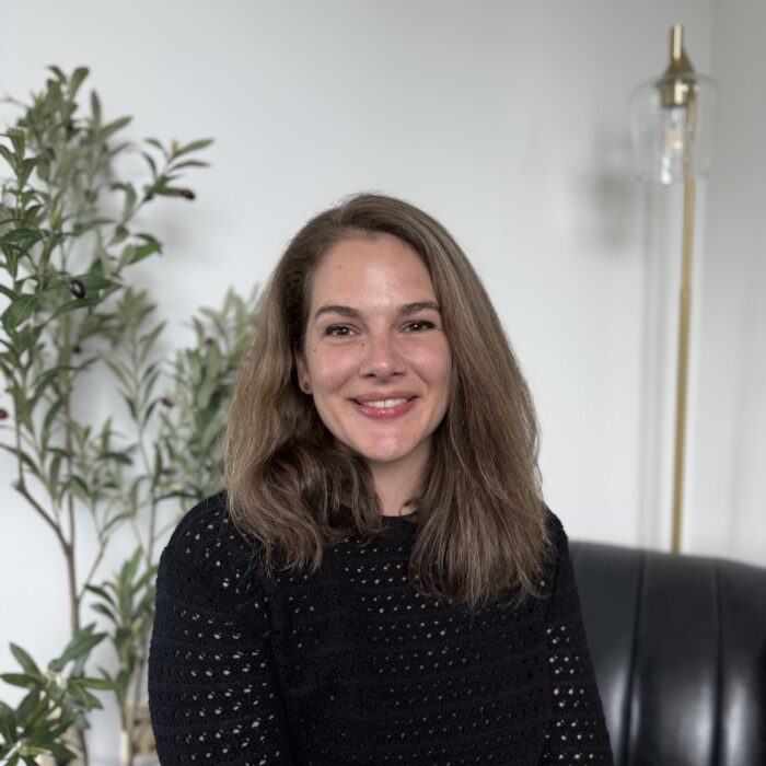 A woman with shoulder-length brown hair and a black sweater smiles while sitting on a black couch. There is a green plant and a modern floor lamp in the background against a white wall.