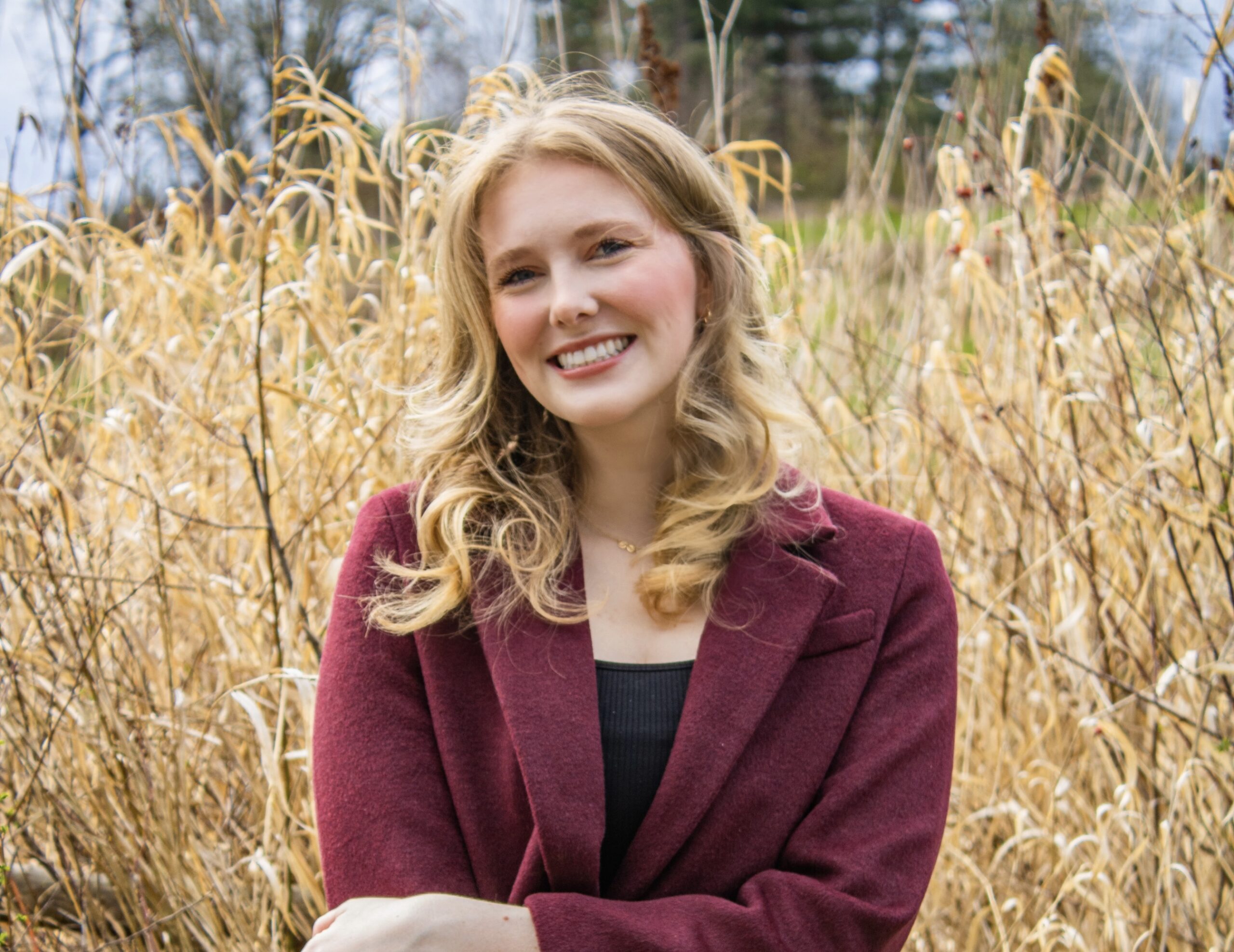A young woman with wavy blonde hair and a maroon blazer smiles while standing in front of tall, dry grass and trees in an outdoor setting.
