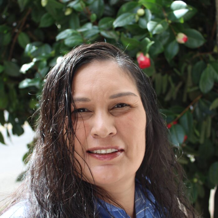 A woman with long dark hair and a blue shirt smiles in front of a leafy green bush with pink flower buds.