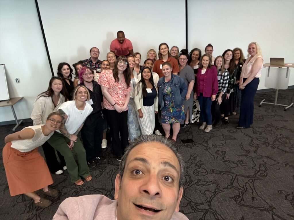 A group of about 25 people pose and smile together in a conference room, while a man in the foreground takes a selfie. The group stands in front of a blank projector screen.