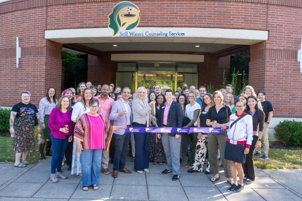 A group of people stands smiling in front of the Still Waters Counseling Services building as two people cut a ceremonial ribbon, celebrating an event. The group appears cheerful and supportive.