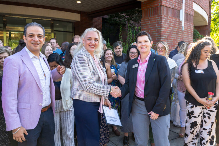 A group of people are gathered outdoors. Two women in the center, dressed in business attire, are shaking hands and smiling at the camera. Others stand around them, some also smiling, in front of a brick building.