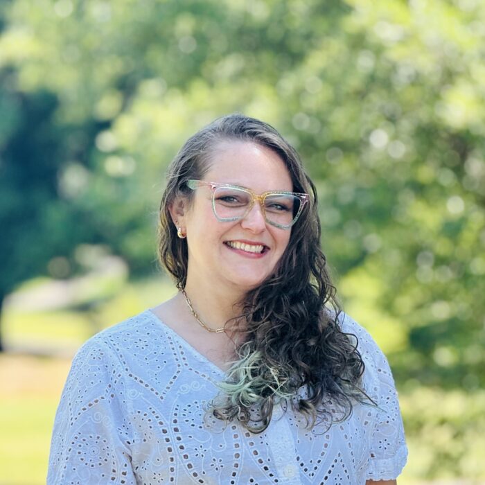 A smiling woman with wavy brown hair wearing glasses and a white eyelet blouse stands outdoors with a green, blurred background of trees and sunlight.
