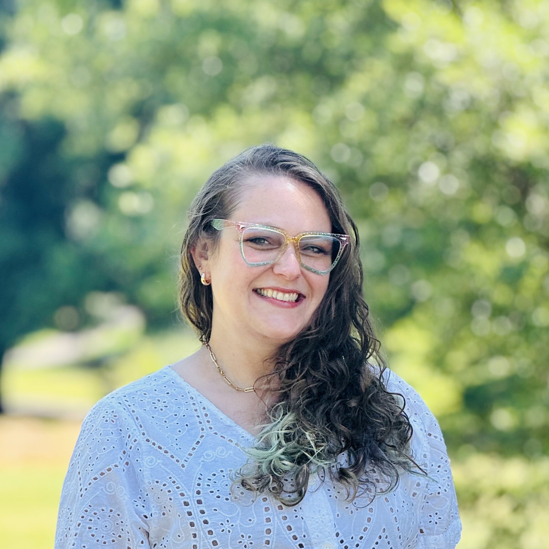 A smiling woman with wavy brown hair wearing glasses and a white eyelet blouse stands outdoors with a green, blurred background of trees and sunlight.