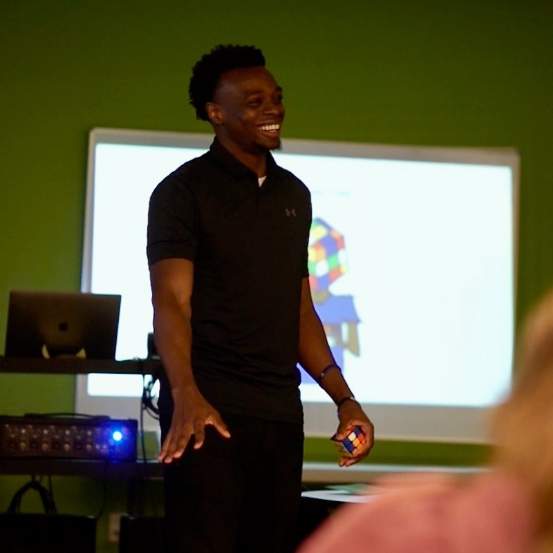 A man in a black shirt stands and smiles in front of a projector screen, engaging with an audience in a classroom or presentation setting. The screen shows a colorful graphic.