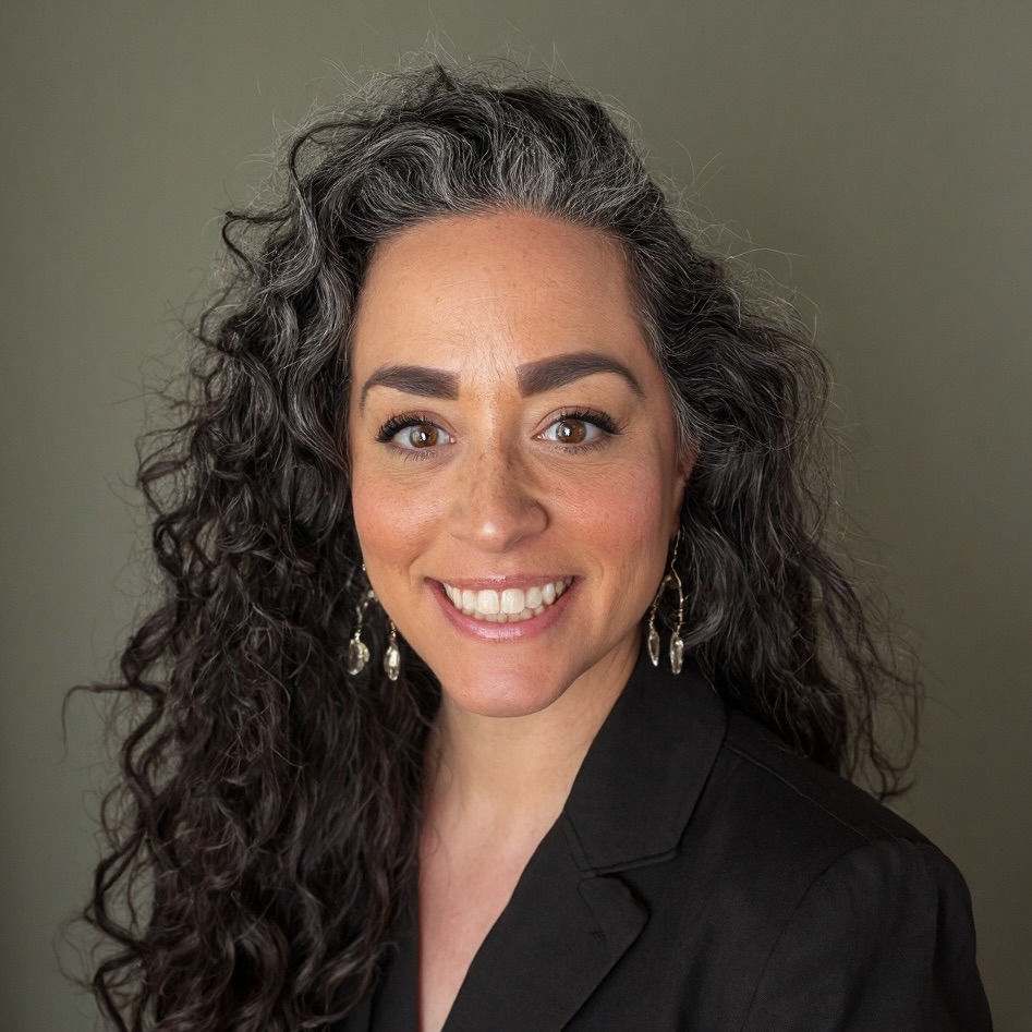A woman with long, curly dark hair with gray streaks, wearing dangling earrings and a black blazer, smiles at the camera against a plain, neutral background.