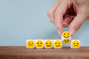 A hand arranges six wooden blocks, each with a different yellow emoji face showing various emotions including sad, angry, confused, neutral, and happy, on a wooden surface against a light blue background.