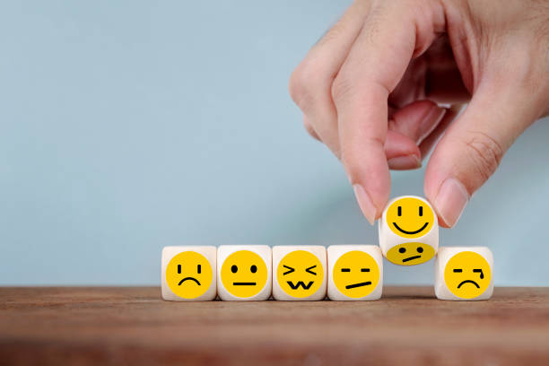 A hand arranges six wooden blocks, each with a different yellow emoji face showing various emotions including sad, angry, confused, neutral, and happy, on a wooden surface against a light blue background.