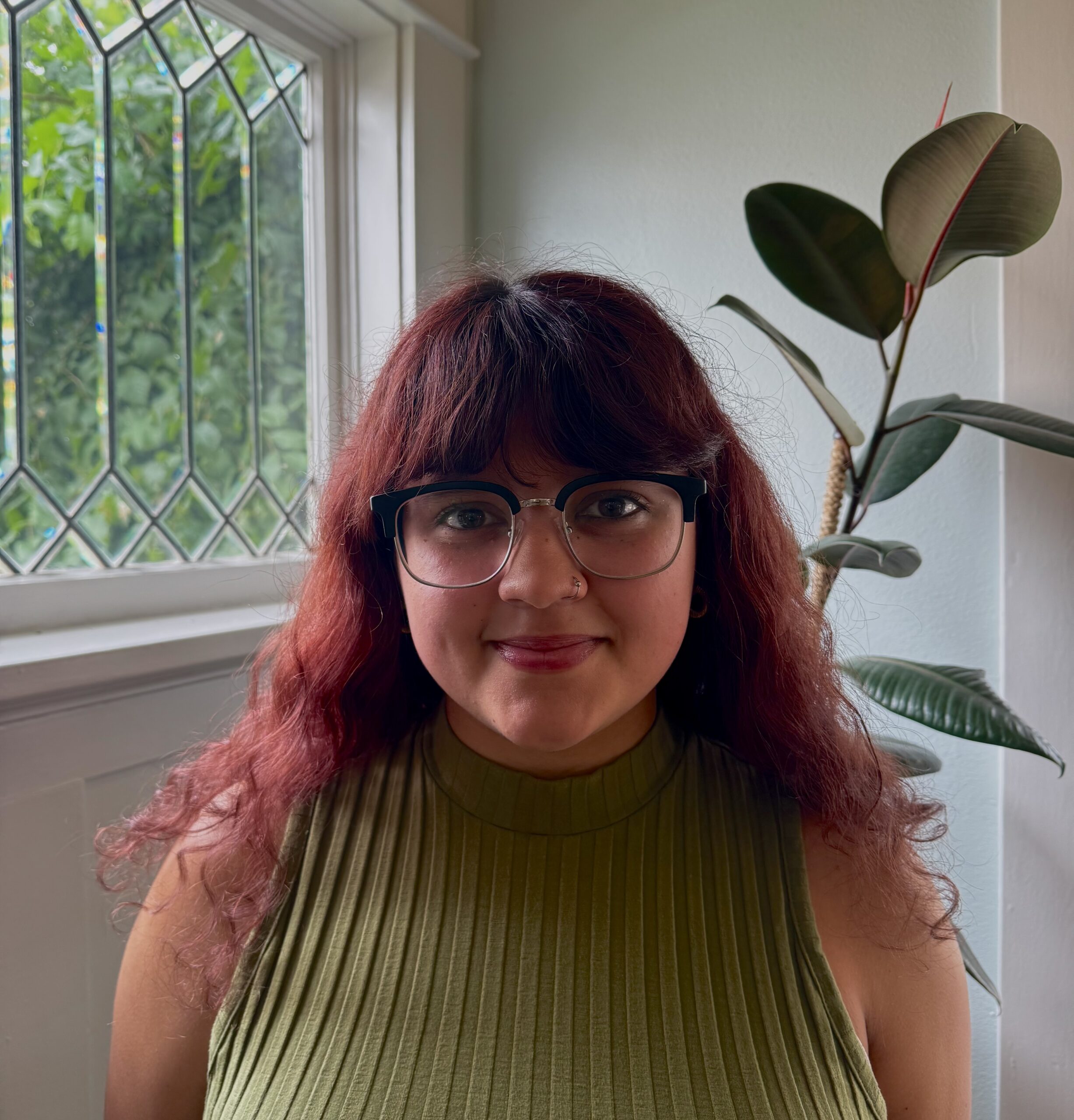 A person with wavy, reddish-brown hair and glasses smiles indoors near a window with decorative glass. They wear a sleeveless, ribbed green top and stand in front of a leafy houseplant.