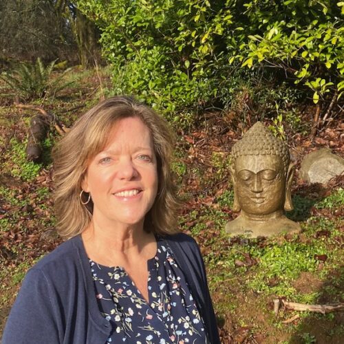 A woman with shoulder-length light brown hair, wearing a navy blue cardigan and floral top, smiles outdoors next to a stone Buddha head statue surrounded by greenery and sunlight.