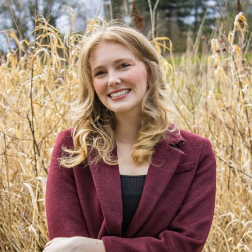 A young woman with wavy blonde hair, wearing a maroon blazer and black top, stands smiling in front of tall, dry grass outdoors on a cloudy day.
