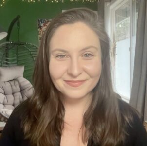 A woman with long brown hair and a black top smiles at the camera in a cozy room with green walls, a hanging chair, string lights, artwork, and a window with curtains in the background.