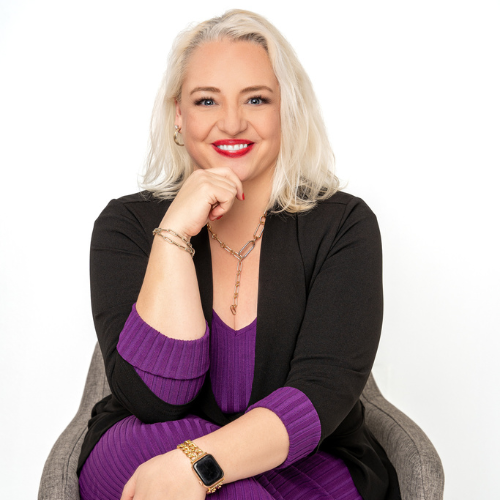 A smiling woman with long, blonde hair sits on a chair, wearing a purple dress and black cardigan. She has her chin resting on one hand and is wearing a smartwatch, necklace, and bracelets. The background is white.