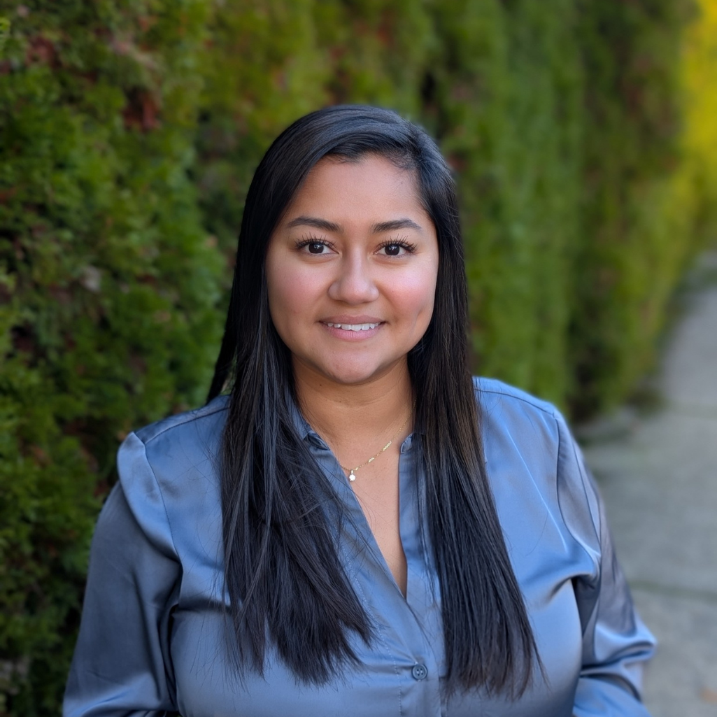 A woman with long dark hair, wearing a gray blouse and a necklace, stands outdoors next to a green hedge and smiles at the camera.