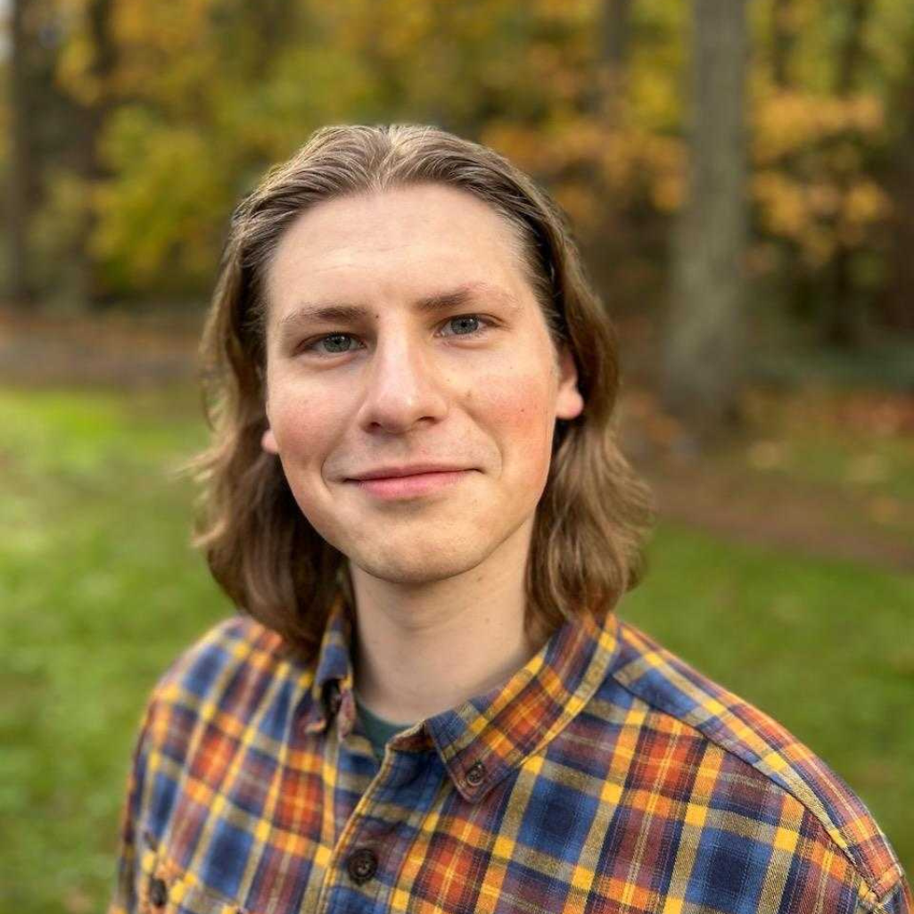 A young man with shoulder-length brown hair smiles outdoors, wearing a yellow and blue plaid shirt. The background is blurred with green grass and tall trees with autumn foliage.