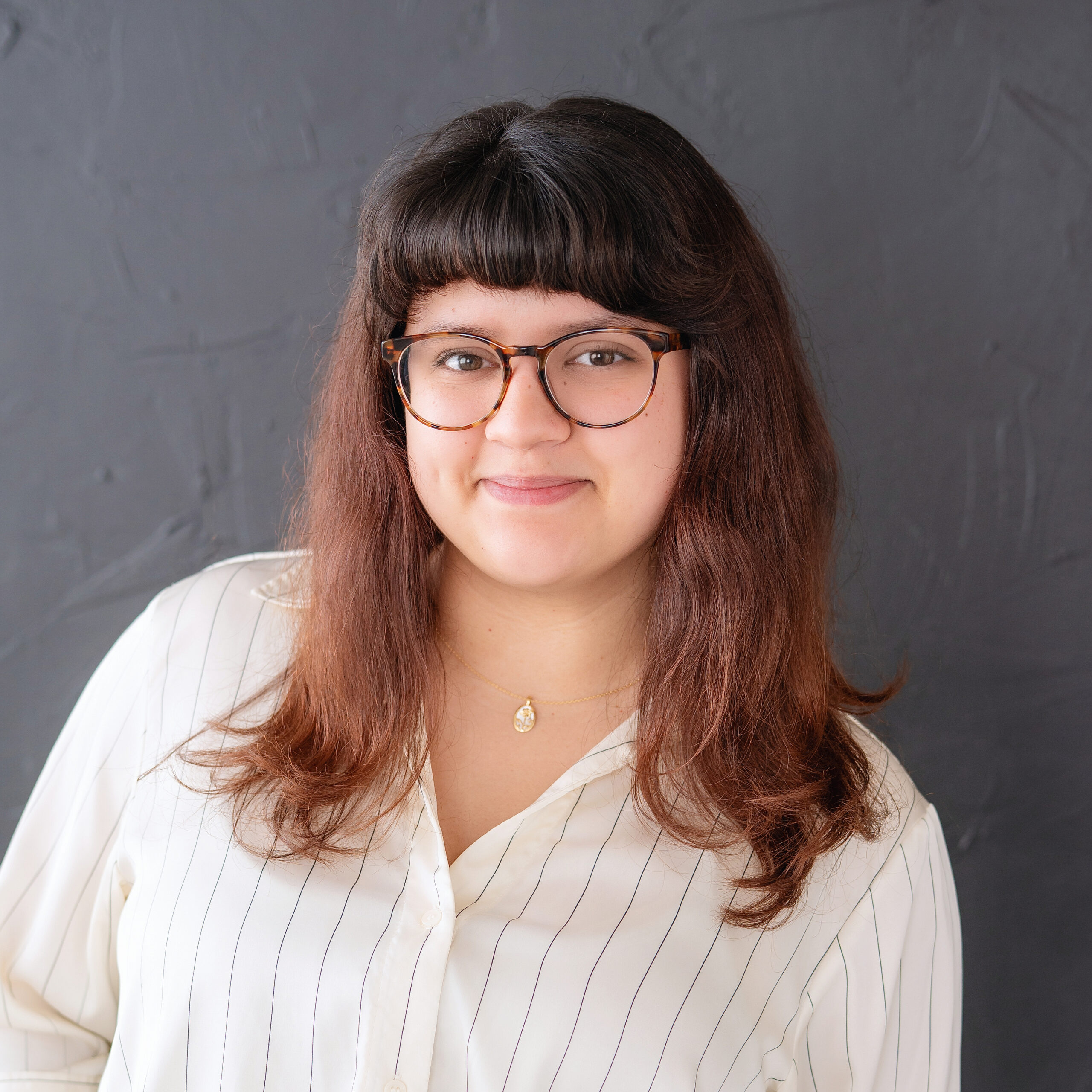 A woman with shoulder-length brown hair and glasses smiles softly. She wears a cream-colored, striped blouse and a gold pendant necklace. The background is a textured, dark gray wall.