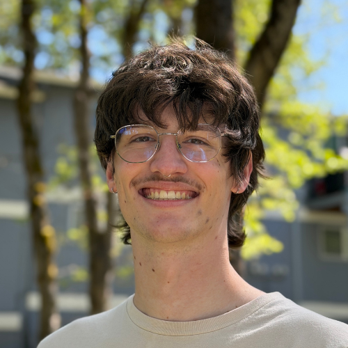 A young man with wavy brown hair, glasses, and a faint mustache smiles outdoors in front of blurred trees and a gray building. He wears a light beige shirt and is in bright sunlight.