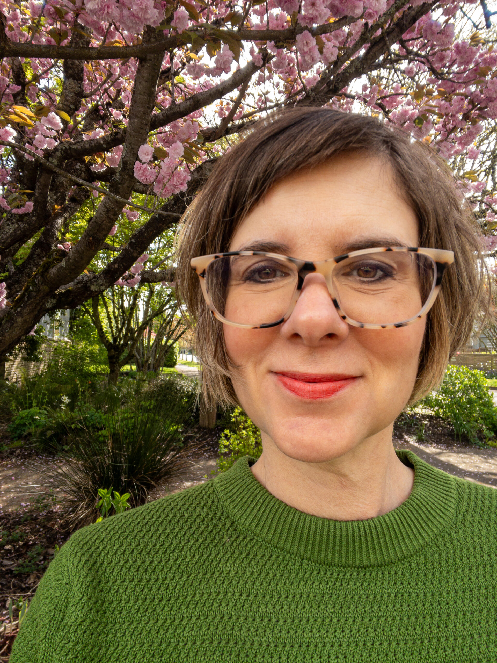 A woman with short brown hair and glasses, wearing a green sweater, smiles outdoors in front of a blossoming tree with pink flowers.