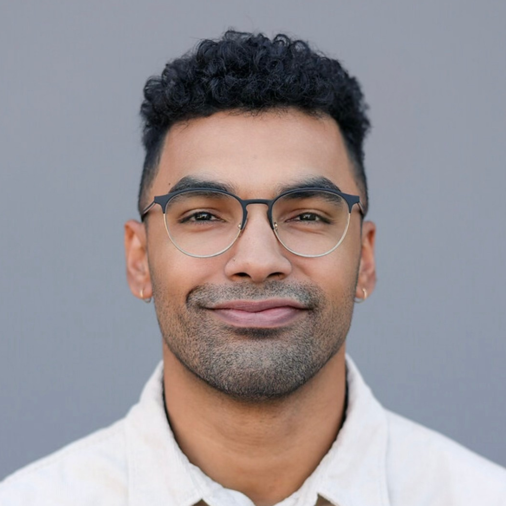 A young man with short curly dark hair, glasses, and small hoop earrings, wearing a light-colored collared shirt, smiles softly in front of a plain gray background.