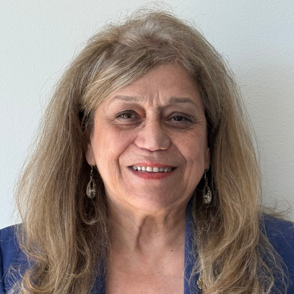 Smiling older woman with long, light brown hair, wearing a blue blazer and dangly earrings, standing in front of a plain light-colored wall.