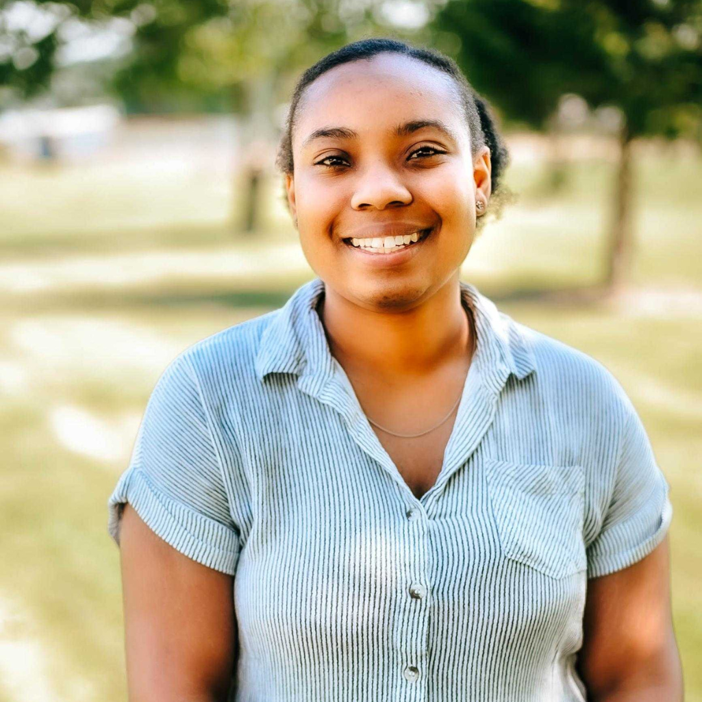 Smiling person standing outdoors in a park, wearing a light blue short-sleeve shirt. Green trees and grass are blurred in the background, creating a bright and pleasant atmosphere.