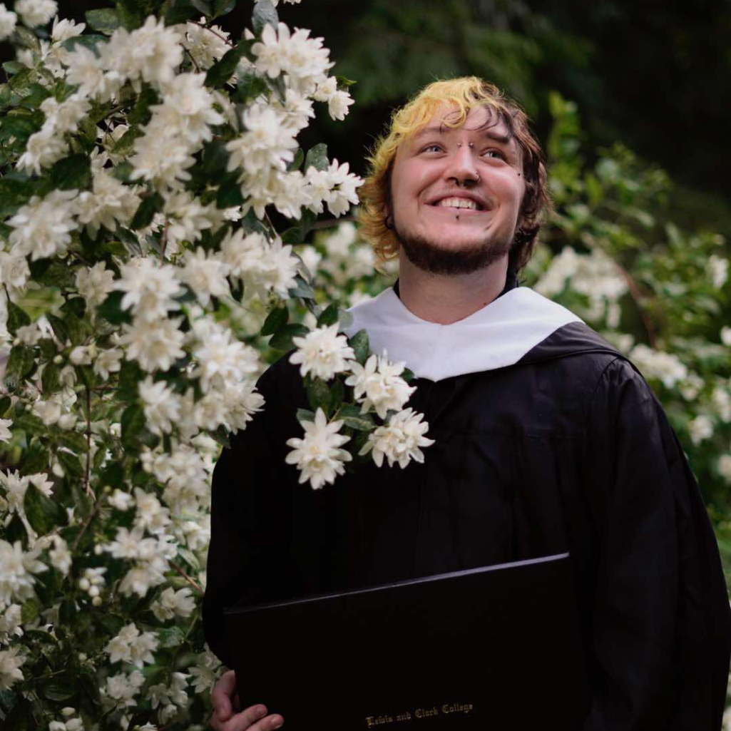 A person in a graduation gown smiles while standing next to blooming white flowers, holding a diploma. Lush greenery fills the background.