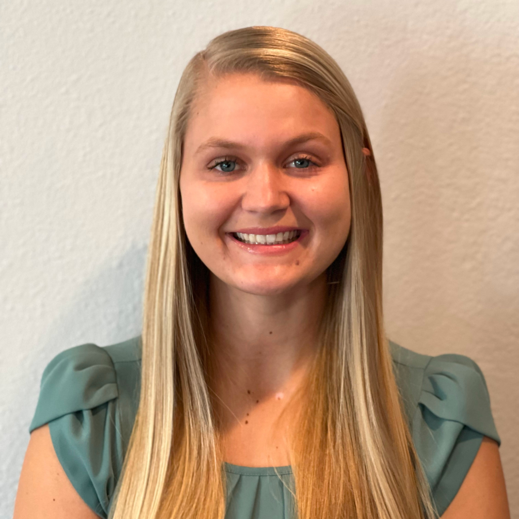 A young woman with long blonde hair, wearing a green blouse, smiles at the camera while standing in front of a light-colored wall.