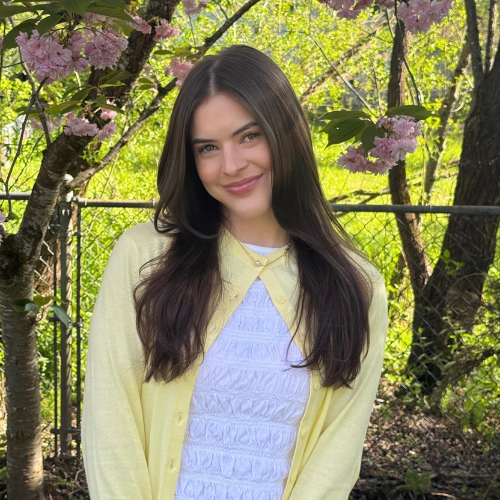 A young woman with long brown hair stands outdoors in front of a tree with pink blossoms, wearing a white top and a light yellow cardigan, smiling at the camera. A chain-link fence and greenery are in the background.