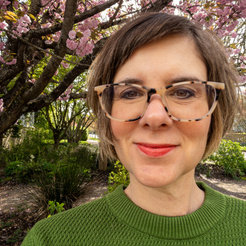 A woman with short brown hair and tortoiseshell glasses smiles outdoors. She wears a green sweater, and blooming pink cherry blossoms and green foliage are visible in the background.