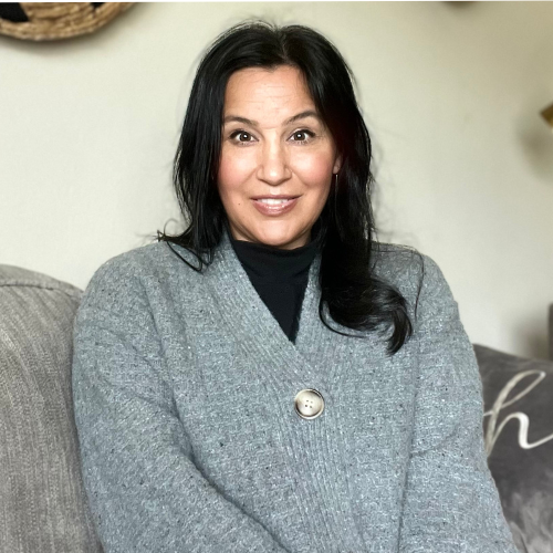 A woman with long dark hair sits on a gray couch, wearing a gray cardigan with a large button and a black top. She is smiling softly at the camera. The background is neutral and softly lit.