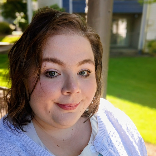 A woman with short brown hair and light skin smiles softly at the camera while standing outdoors on a sunny day, with green grass and a building visible in the background.