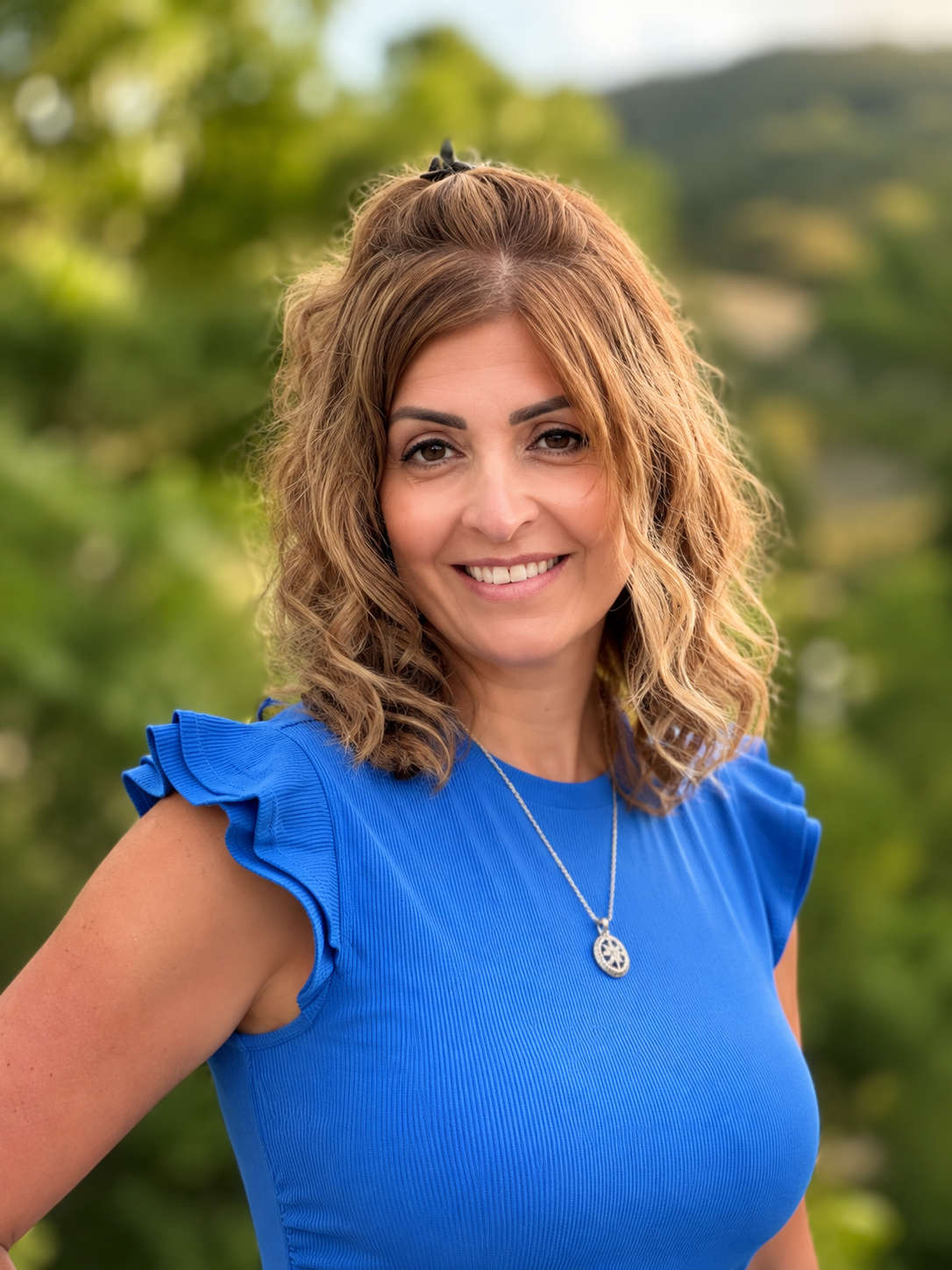 A woman with wavy, shoulder-length hair and a blue ruffled top smiles outdoors, wearing a silver necklace, with green trees and hills blurred in the background.