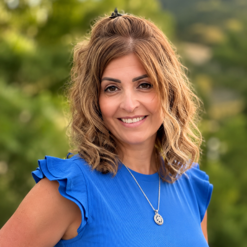 A woman with wavy light brown hair, wearing a bright blue ruffled top and a necklace, smiles outdoors with green trees in the blurred background.