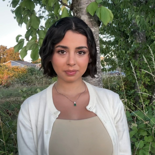A woman with short dark hair, wearing a white cardigan and a pendant necklace, stands outdoors in front of green trees and foliage, with houses visible in the background.