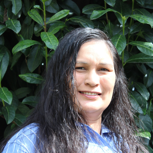 A woman with long dark hair and a blue shirt smiles outdoors in front of lush green leaves.