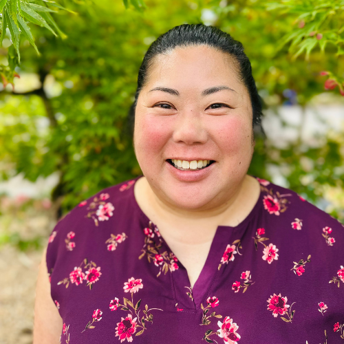 A woman with dark hair in a bun, wearing a purple floral top, smiles outdoors with green leaves blurred in the background.