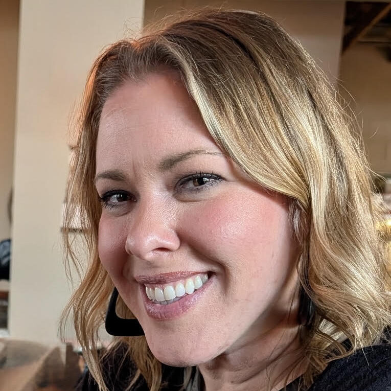 A woman with wavy blonde hair, wearing a black top and earrings, smiles warmly indoors with blurred background elements.